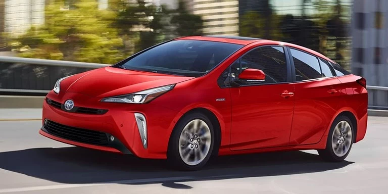 A bright red Toyota Prius, an electric vehicle widely available in the New Zealand market, features a sleek design and an aerodynamic structure, with a hatchback shape and low roofline