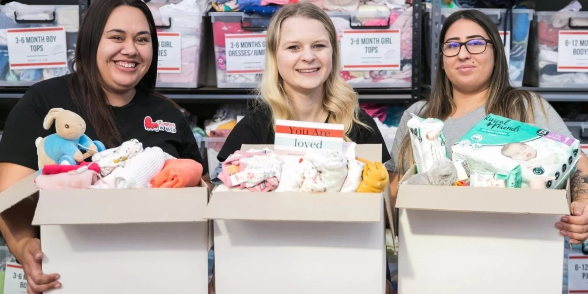 Three women with boxes full of donated baby clothes, toys, and other baby items posing for a photo at Mummy's in Need