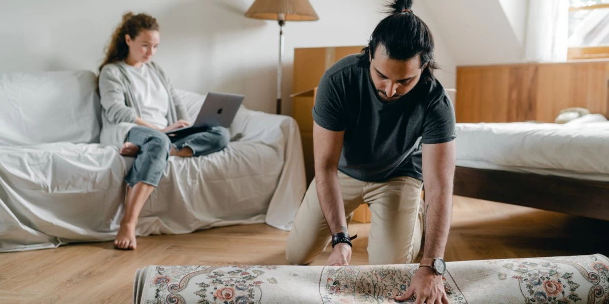 A man unrolling a rug onto the floor of his new rental place, a simple, budget-friendly way to instantly bring comfort and style to his home.