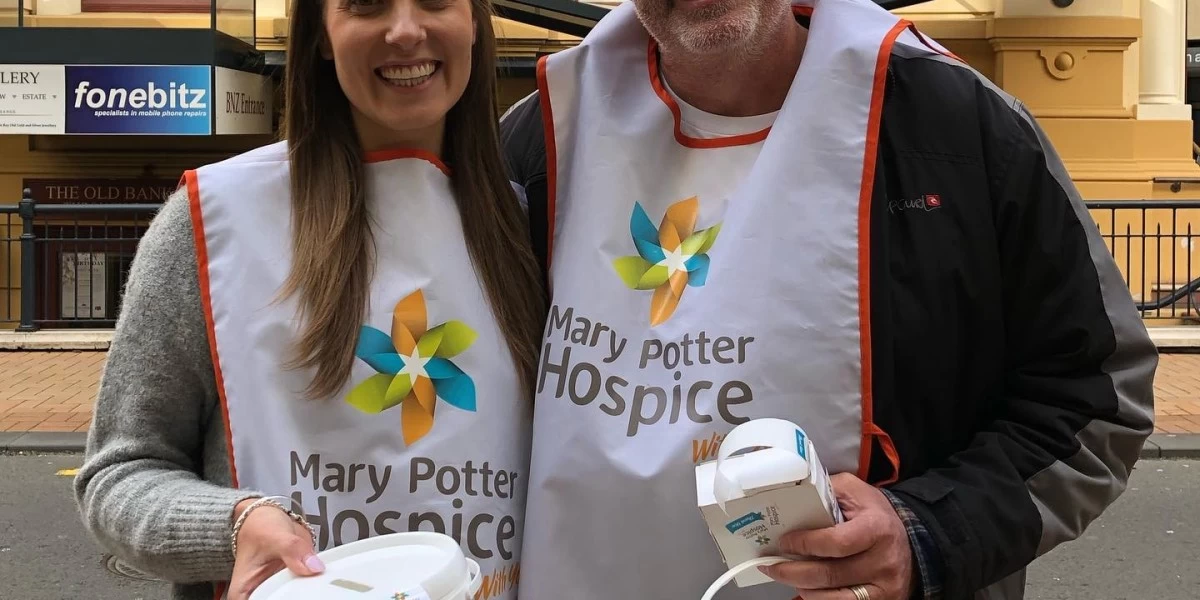 A man and a woman with wearing hospice aprons posing for a photo, holding items from a Hospice shop