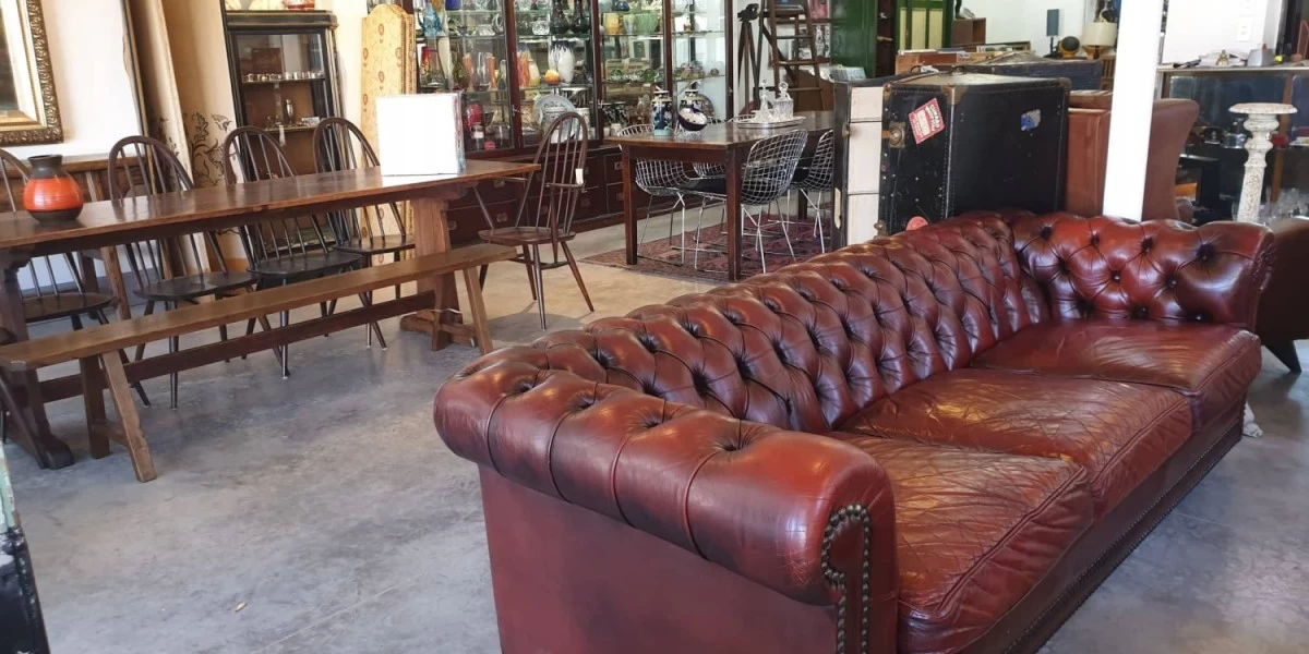 A vintage brown leather couch, with vintage dining tables and chairs in the background, being sold at Wakefield Antiques store in Greytown