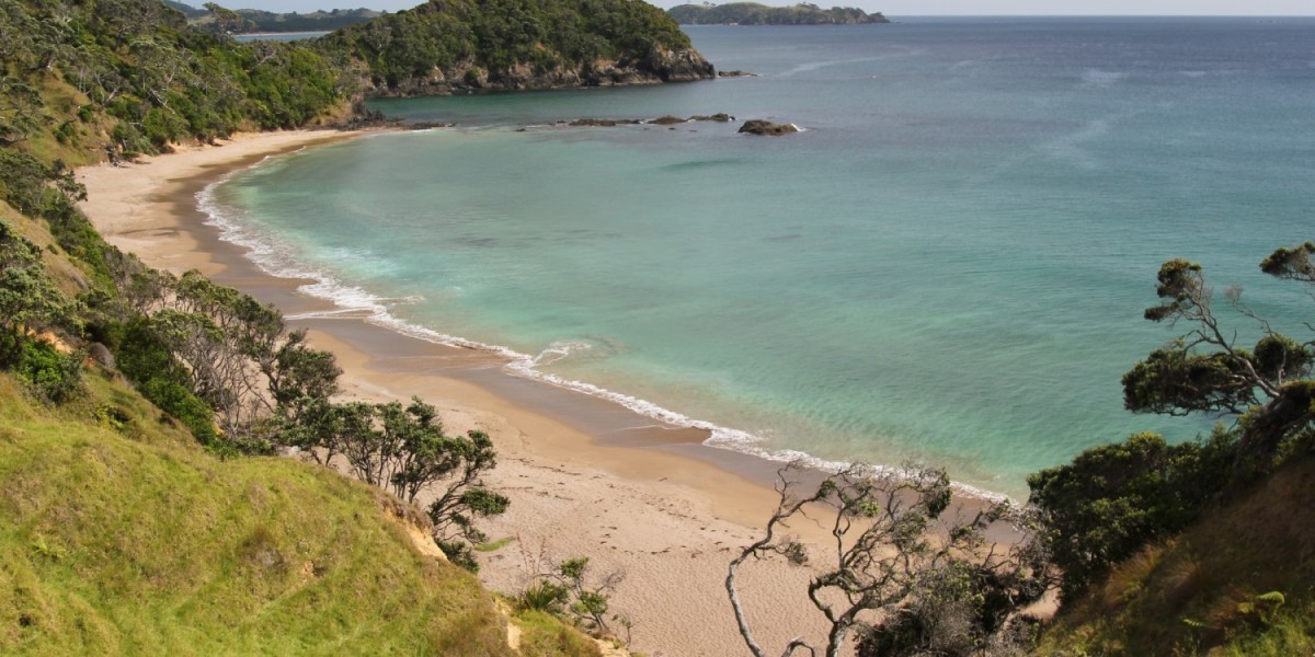 A wide, panoramic view of the vibrant blue Smugglers Bay off the coast of New Zealand, with golden sand and rocky cliffs on either side and scattered islands in the distance.