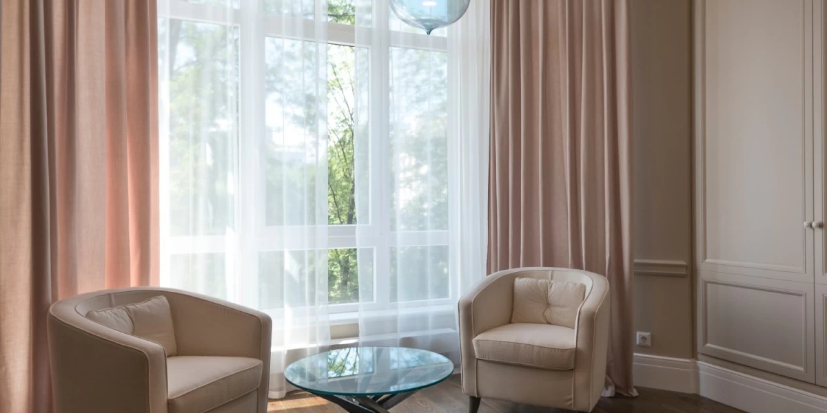 A mid-sized living with beige and sheer white curtains opens up to a bright window, allowing for a gentle glow to come through, reminding one of the feelings of home.
