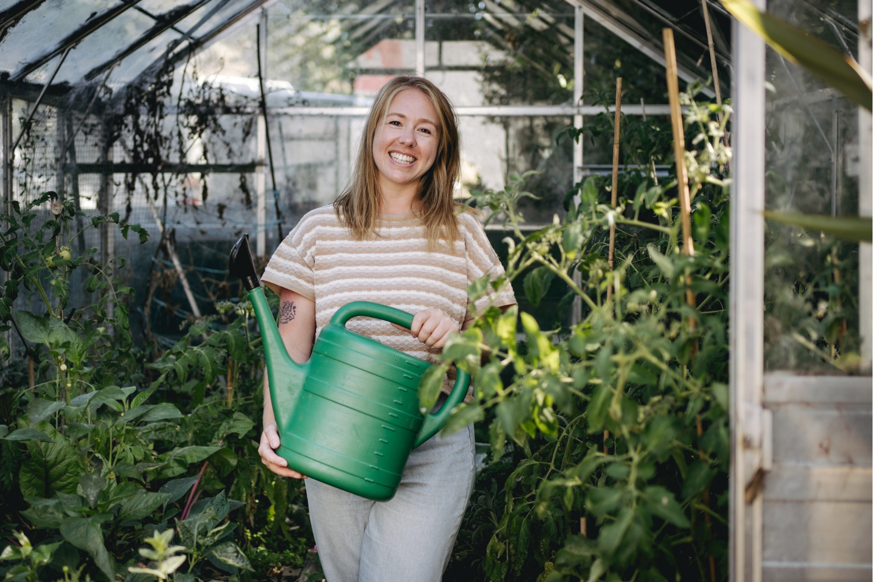 Woman holding a watering can in a greenhouse full of vegetable plants.