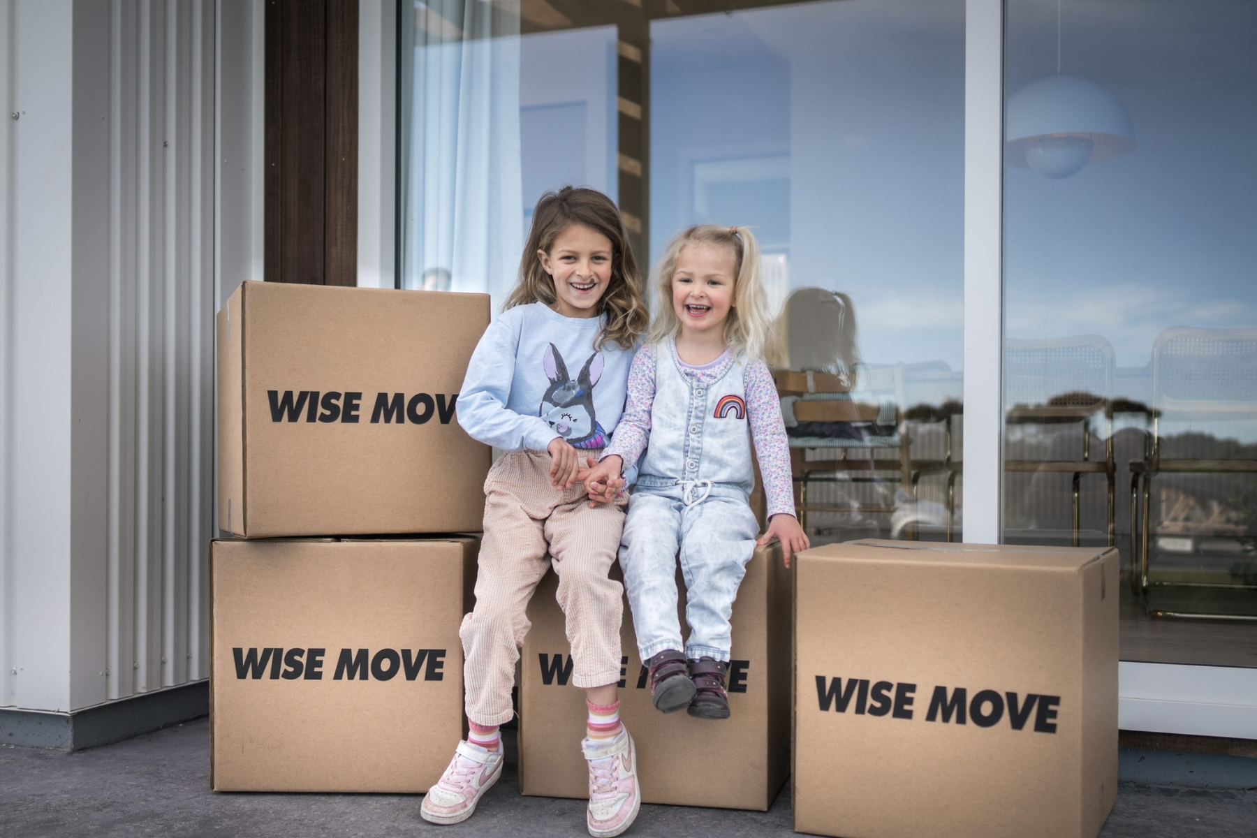 Two children sitting on Wise Move boxes outside a New Zealand home on moving day